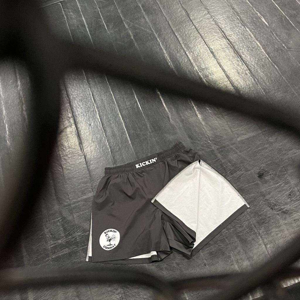 Kickin' Combat "Origins" high cut gladiator MMA shorts on floor behind cage fence