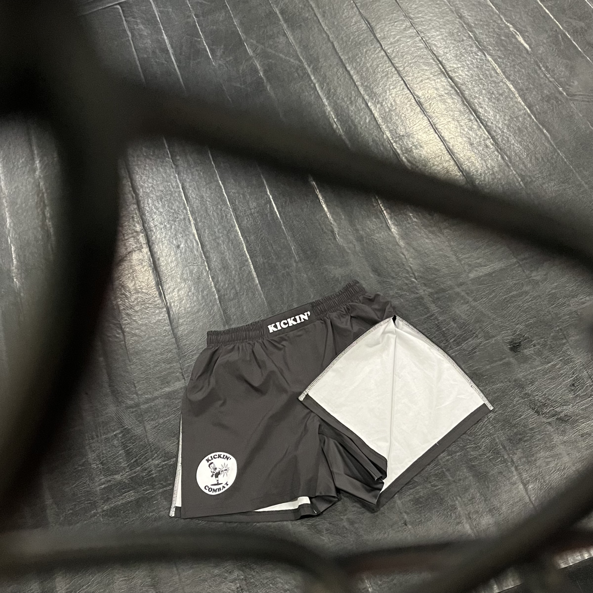 Kickin' Combat "Origins" high cut gladiator MMA shorts on floor behind cage fence