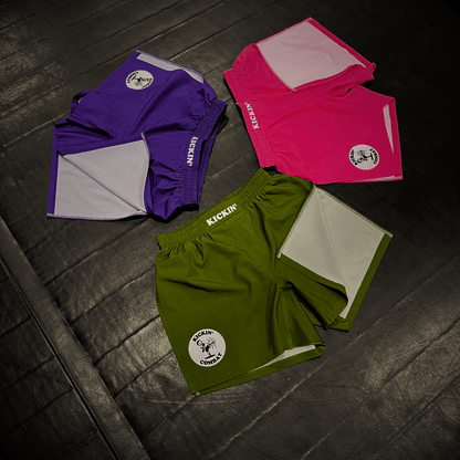 Kickin' Combat "Origins" olive green, hot pink, and purple high cut gladiator MMA shorts folded on fight gym mat