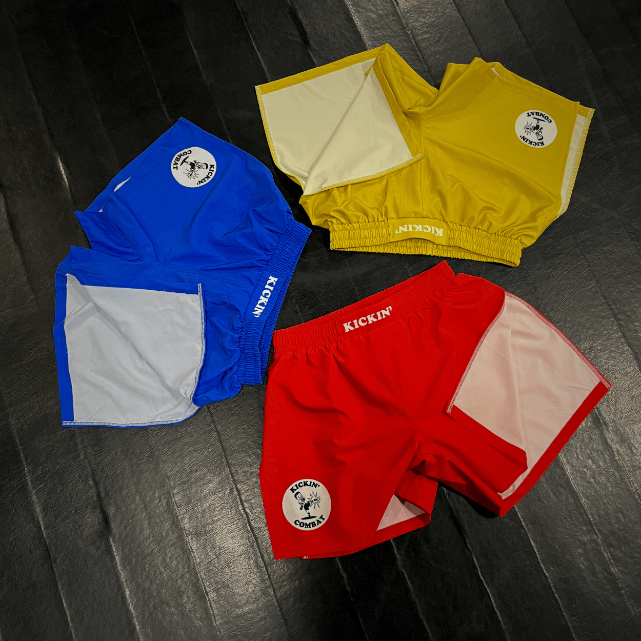 Kickin' Combat "Origins" red, blue, and yellow high cut gladiator MMA shorts folded on fight gym mat